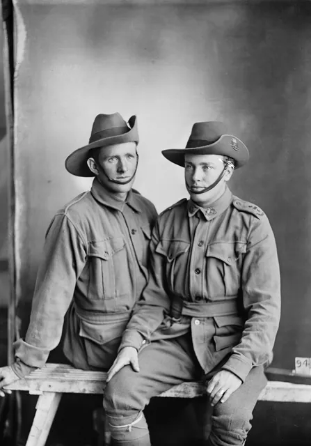 Two young soldiers in slouch hats and tunics sitting side by side on a rough bench in a photographer’s studio, shoulders touching: the one on the left looks straight at the camera, the one on the right looks away.